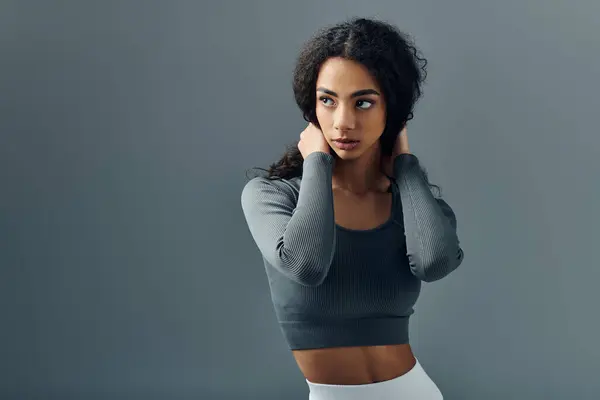 Stock image A young woman in a gray top stands in front of a gray background, looking to the side and adjusting her hair.