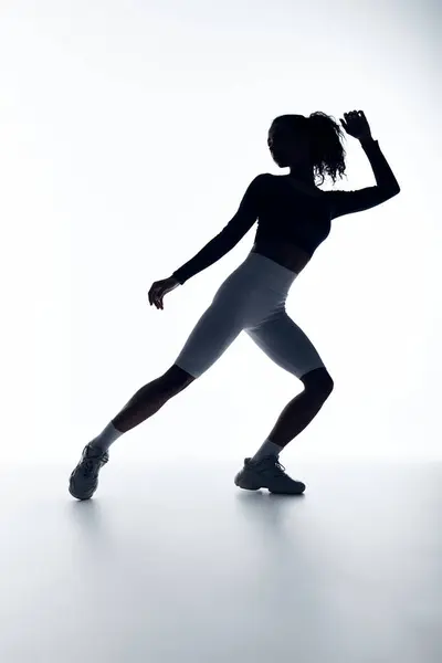 stock image A young woman in athletic wear stretches her leg, silhouetted against a white background.