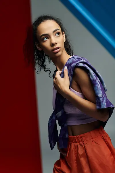 stock image A young woman in a sports bra and orange shorts leans against a red wall in a studio setting, with colorful glass panels in the background.