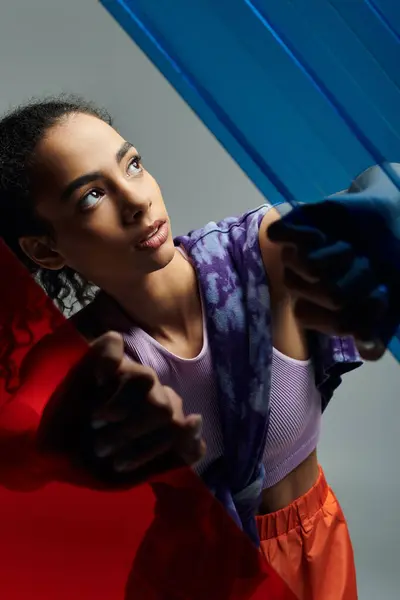 stock image A young African American woman in athletic wear stretches towards colorful glass panels in a studio, her eyes gazing upwards.