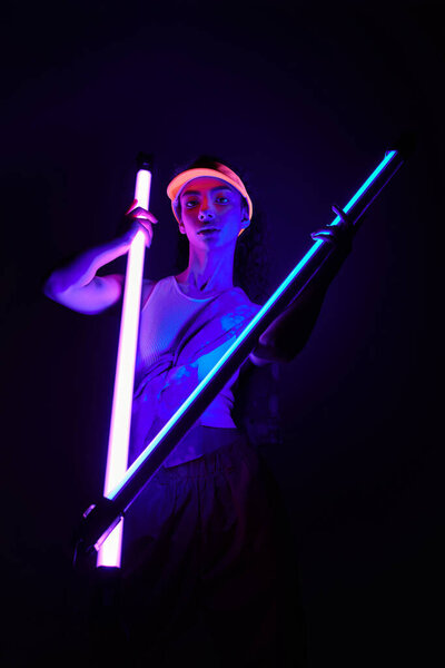 Молодая чернокожая женщина позирует в студии с неоновыми огнями, в непринужденном, но стильном наряде.