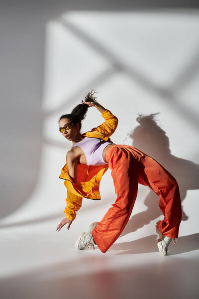 Woman in yellow jacket and orange pants poses intently in studio with gray background, holding her hair back with one hand.