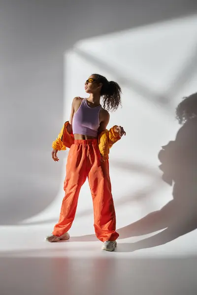 stock image A young woman in a studio wears a yellow jacket, purple tank top, orange pants, and sunglasses, exuding coolness.