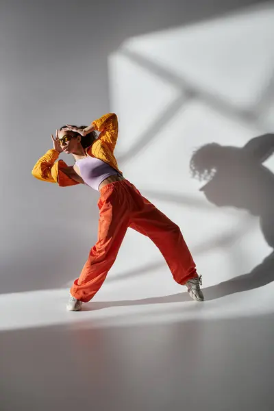 stock image A stylish young woman in yellow jacket and orange pants strikes a confident pose against a gray backdrop, embodying modern fashion.