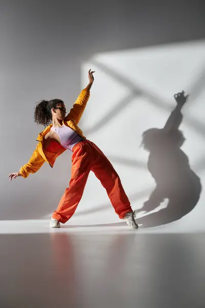 stock image A girl in yellow jacket and orange pants dramatically poses against gray backdrop, shadow extending across studio.