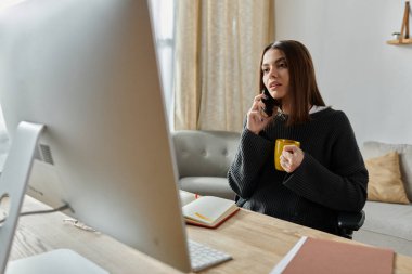 Gri kazaklı genç bir kadın evindeki bir masada oturmuş telefonla konuşuyor ve bilgisayarını tamir ediyor..