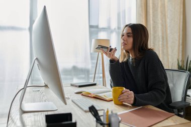 Gri kazaklı genç bir kadın ofis masasında oturuyor. Ağzına telefon dayayıp bir ses kaydediyor..