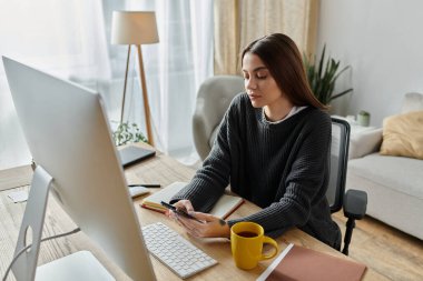Gri kazaklı genç bir kadın masasında oturuyor, telefonuna odaklanmış, yanında bilgisayar monitörü ve diğer iş materyalleri var..