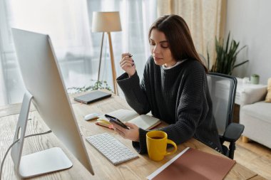 Evde çalışan genç bir kadın telefonuna odaklanırken bilgisayar monitörü ve sıcak bir fincan kahve masasında oturuyor..