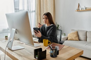 Genç bir kadın bir ofis masasında çalışıyor, telefonunu ve büyük bir monitörü kullanıyor..