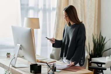 A young woman in a grey sweater works at her home office desk, looking at her phone and holding paperwork. clipart