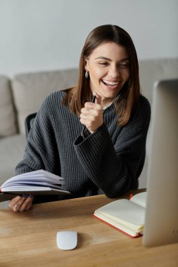 A young woman in a grey sweater sits at her desk, smiling as she works on her computer and takes notes in her journal. clipart