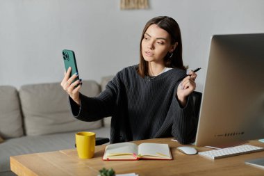 Genç bir kadın masasında oturuyor, telefonuna bir video kaydediyor, düşüncesizce diğer elinde bir kalem tutuyor..