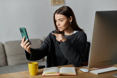 Gri kazaklı genç bir kadın evinde masasında otururken telefonunda içerik yaratıyor..