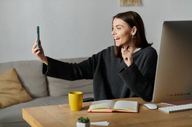 Gri kazak giyen genç bir kadın, bir ofis masasında oturuyor. Telefonuyla bir video kaydediyor ve gülümsüyor..
