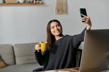 Genç bir kadın ofisteki bir masada oturuyor. Elinde fincanı tutarken telefonuyla selfie çekiyor..