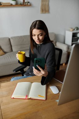 Genç bir kadın masasında oturuyor, uzaktan çalışıyor. Bir telefon tutuyor, içerik yaratıyor ve kahvesini yudumluyor..