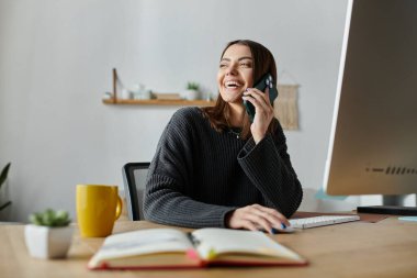 Gri kazaklı genç bir kadın masasının önünde elinde bir not defteri ve kahve fincanıyla telefonla konuşurken gülümsüyor..