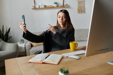 Gri kazaklı genç bir kadın, ofisteki bir masada otururken telefonundan içeriğini filme alıyor..