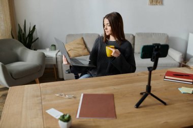 A young woman in a grey sweater sits at a table, working on a laptop and recording a video for her followers. clipart