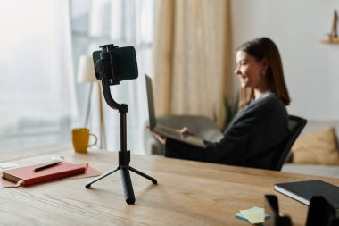 Arka planda dizüstü bilgisayarda çalışan genç bir bayanla birlikte tripoda akıllı bir telefon yerleştiriliyor..