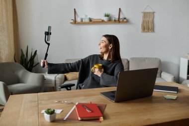 Gri kazaklı genç bir kadın elinde kahve fincanı tutarken gülümsüyor ve bir ofis masasında telefonuyla video kaydediyor..