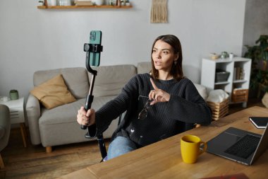 Gri kazak giyen genç bir kadın, ev ofisinin masasında oturmuş, selfie çubuğu ve akıllı telefon kullanarak içeriği kaydediyor..