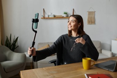 Gri süveter giyen genç bir kadın, özçekim çubuğuna bağlı akıllı bir telefonla içeriğini çekerken gülümsüyor..