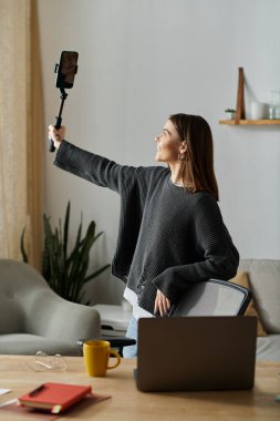 Gri kazaklı genç bir kadın evde uzaktan çalışırken sosyal medya için içeriği filme alıyor..