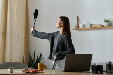 Gri kazaklı genç bir kadın, ofis masasında beklerken akıllı telefon ve selfie çubuğuyla içeriğini filme alıyor..