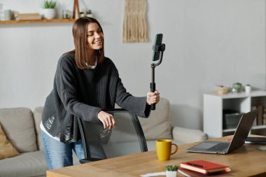 Gri kazaklı, akıllı telefonlu, selfie 'li genç bir kadın masadaki bir koltuğun arkasında dizüstü bilgisayarla oturuyor..