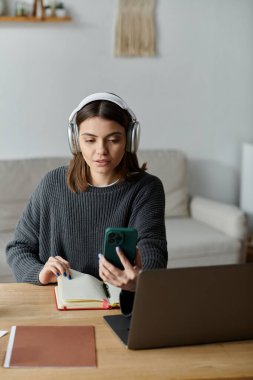 Gri kazaklı genç bir kadın masasında oturuyor, kulaklıkları takılı ve telefonuna dikkatlice bakıyor, mesleğinin araçlarıyla çevrili..