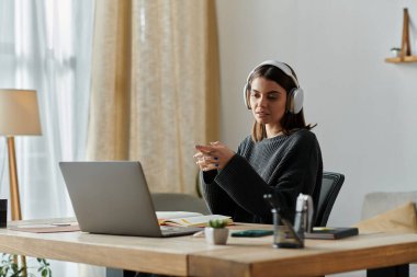 A young woman in a grey sweater sits at a desk in her home office, working on a laptop while wearing headphones. clipart