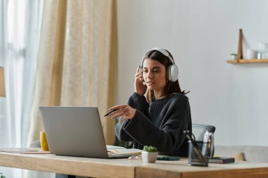 A young woman sits at a desk, wearing headphones, and working on a laptop computer. She is creating content for her business. clipart