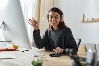 Kulaklık takan genç bir kadın ofis masasında çalışırken gülümsüyor ve jestler yapıyor..