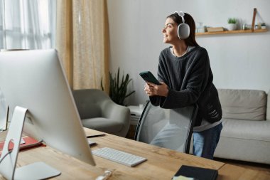 Gri kazaklı bir kadın evden uzaktan çalışıyor. Telefonunu ve bilgisayarını kullanırken gülümsüyor..