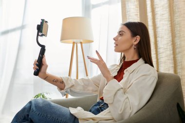 Oturma odasında akıllı telefon dengeleyicisiyle video kaset çeken genç bir kadın. Rastgele giyinerek, seyircileriyle takılır..