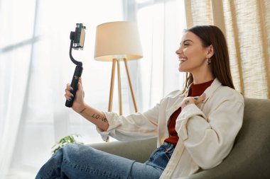 Genç bir kadın, sıradan giyinmiş, bir kanepede oturuyor ve elinde akıllı telefon sabitleyicisi tutarken video kaydediyor..