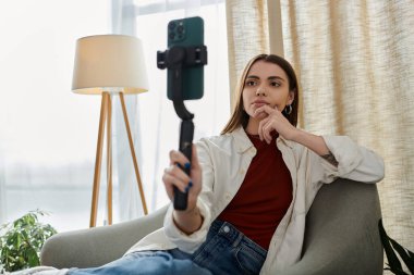 Oturma odasında oturan genç bir kadın, sıradan giyinmiş ve akıllı telefon dengeleyicisi kullanarak video kaydediyor..