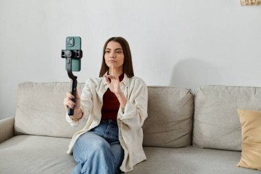Gündelik kıyafetli genç bir kadın kanepeye oturur ve akıllı telefon dengeleyicisi kullanarak video çeker..