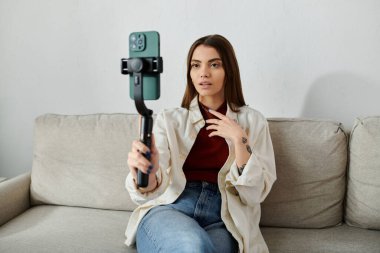 Günlük giysiler içinde bir kadın kanepeye oturup video bloğu çekiyor ve akıllı telefon dengeleyicisiyle dijital pazarlama becerilerini gösteriyor..