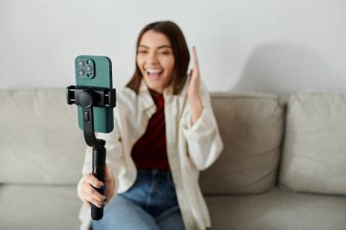 Gülümseyen genç bir kadın kanepede otururken akıllı telefon dengeleyicisi kullanarak bir video bloğu çekiyor..