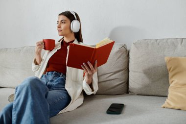 Gündelik kıyafetli genç bir kadın, elinde kitap tutarken ve bir fincandan yudumlarken kulaklıkla müzik dinliyor..