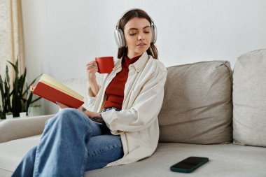 Günlük giysiler ve kulaklıklar takan genç bir kadın elinde kitap ve fincanla bir kanepede dinleniyor..