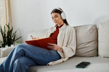 Kulaklık takan genç bir kadın oturma odasındaki kanepede oturup kırmızı bir deftere yazı yazıyor..