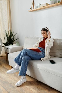 Günlük kıyafetler giyen genç bir kadın oturma odasındaki beyaz bir kanepede dinleniyor, kablosuz kulaklıkla müzik dinliyor..