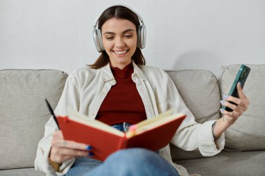 Günlük kıyafetli genç bir kadın kanepede dinleniyor, kablosuz kulaklıkla müzik dinliyor ve kırmızı bir kitap okuyor..