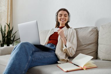 Kulaklık takan genç bir kadın dizüstü bilgisayarı ve not defteriyle koltukta oturuyor, evde çalışırken gülümsüyor..
