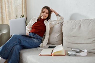 Genç bir kadın dizüstü bilgisayarla evden uzaktan çalışıyor, yanında bir not defteri ve kulaklıkla rahatça oturuyor..