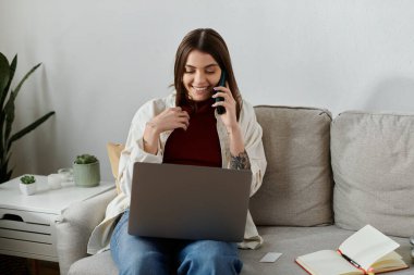 Genç bir kadın kanepede oturuyor, dizüstü bilgisayar kullanıyor ve evde çalışırken telefonla konuşuyor..